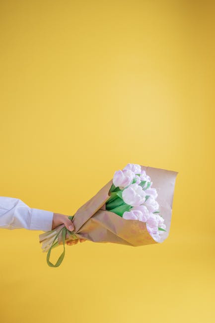 A person dressed in a white shirt is holding a bouquet of pastel pink and white tulips, wrapped in light brown paper tied with a green ribbon, against a vibrant yellow background. The tulips have fres
