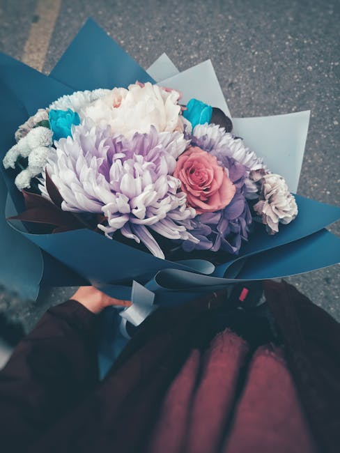 A bouquet of fresh flowers held by a person, featuring a large white chrysanthemum, soft pink rose, purple statice, and smaller blue and white accent flowers, all arranged in a harmonious display. The