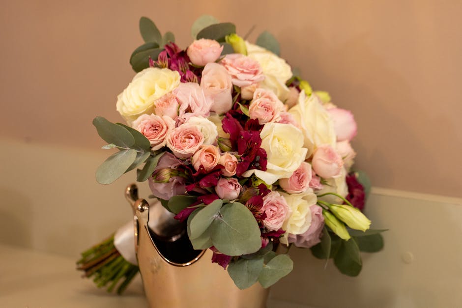 A bouquet of fresh flowers arranged in a compact, rounded style featuring pale pink and white roses, complemented by dark red ranunculus and lisianthus, accented with lush eucalyptus leaves. The bouqu
