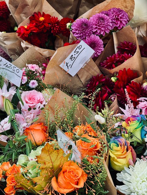 A vibrant assortment of freshly cut flowers arranged in small brown paper packaging, featuring red dahlias, pink and purple chrysanthemums, orange roses with green foliage, and rainbow-colored roses. 