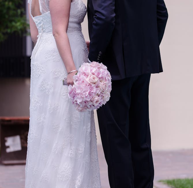 A bride and groom stand close together, with the bride wearing a white lace wedding gown and the groom in a dark suit. The bride holds a round bouquet of soft pink roses and pale pink hydrangeas, whic