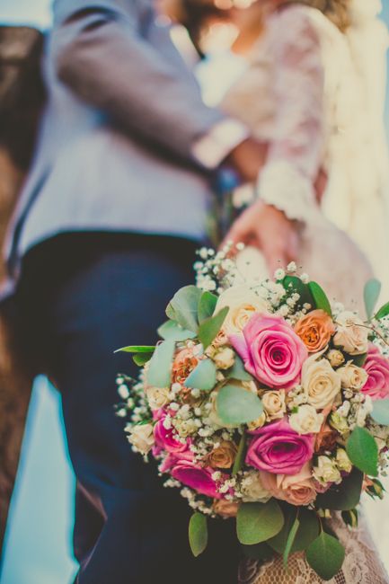 A close-up of a vibrant floral bouquet held by a person, comprising pink roses, cream-colored roses, small peach flowers, and sprigs of baby's breath, accented with green eucalyptus leaves. The bouque