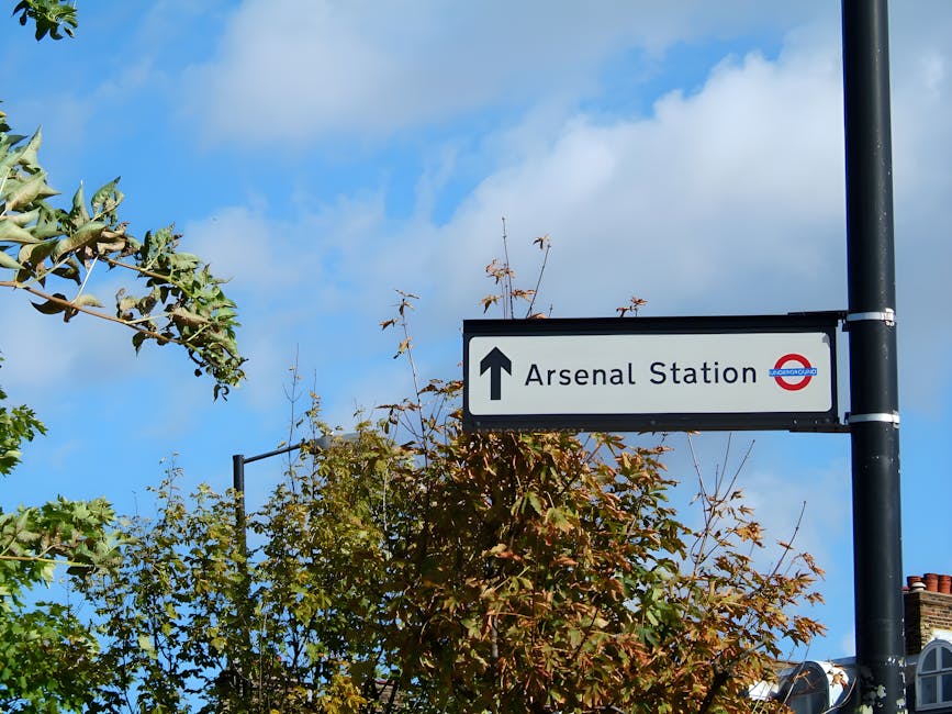 A street sign indicating the direction to Arsenal Station, mounted on a tall black pole, surrounded by leafy trees with green and reddish-brown leaves. The sign features a white background with black